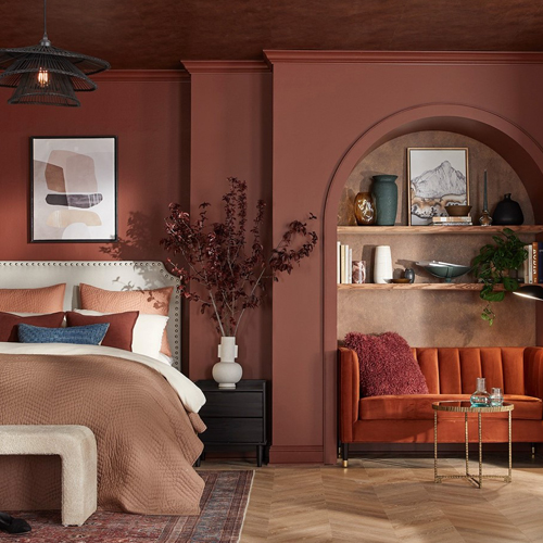 Cozy bedroom with brown walls, a beige upholstered bed with layered pillows and blanket, a black nightstand with a white vase holding dried branches, and an adjacent arch nook featuring a rust-colored sofa with a throw pillow, shelves with decor, and a small round glass coffee table.